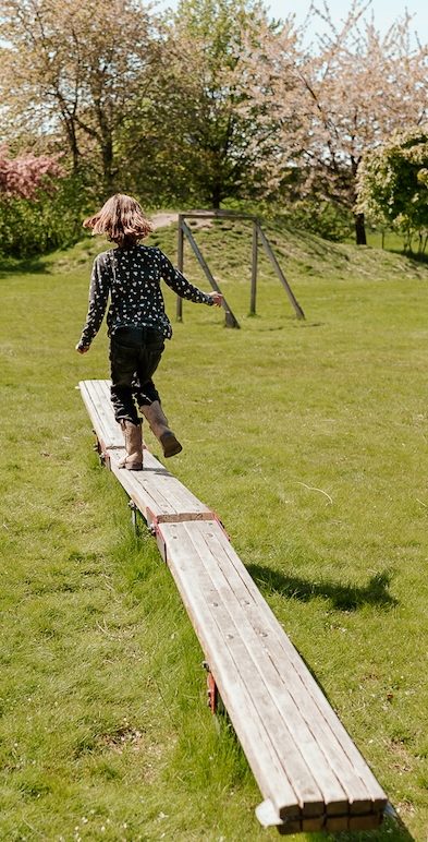 Barn balanserar på en träplanka i förskolans trädgården med blommande träd i bakgrunden.